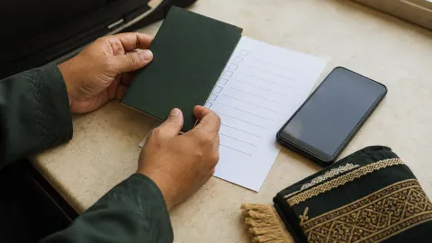 Tangan memegang paspor di samping ceklis dokumen, ponsel, dan sajadah